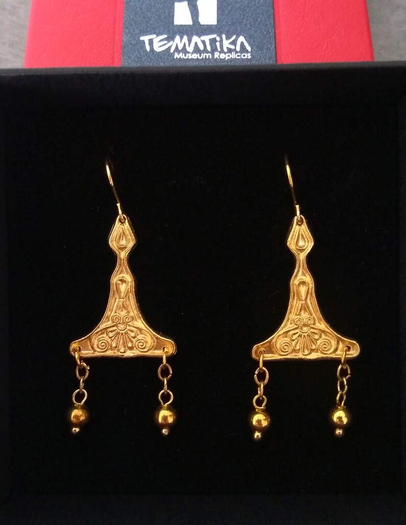 Puede incluir: Un par de pendientes de oro con un dise&ntilde;o triangular y cuentas colgantes. Los pendientes est&aacute;n en una caja negra con el texto "Tematika Museum Replicas".