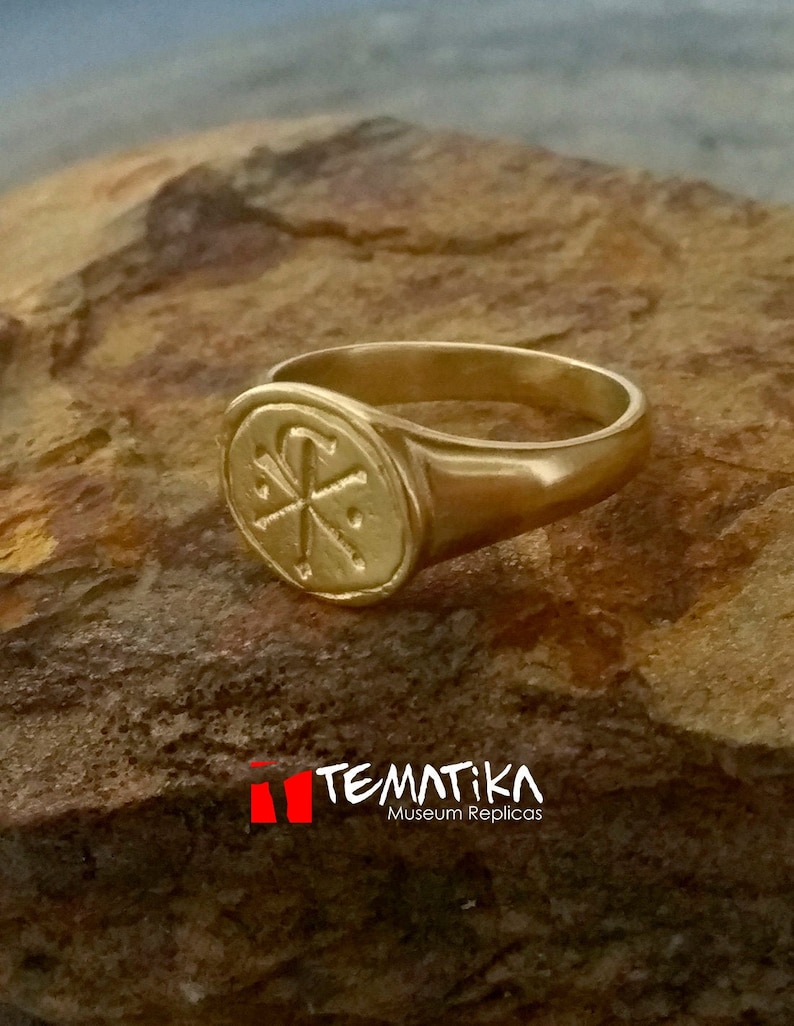 Puede incluir: Un anillo dorado con una cara ovalada con un s&iacute;mbolo Chi Rho. El anillo se muestra sobre una superficie de piedra marr&oacute;n texturizada. La imagen incluye el texto "TEMATIKA Museum Replicas".