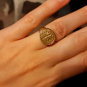 Puede incluir: Un anillo dorado con una cara circular que presenta una cruz y otros s&iacute;mbolos. El anillo se lleva en un dedo, con la mano en primer plano. El dise&ntilde;o del anillo es detallado y parece ser vintage o antiguo.