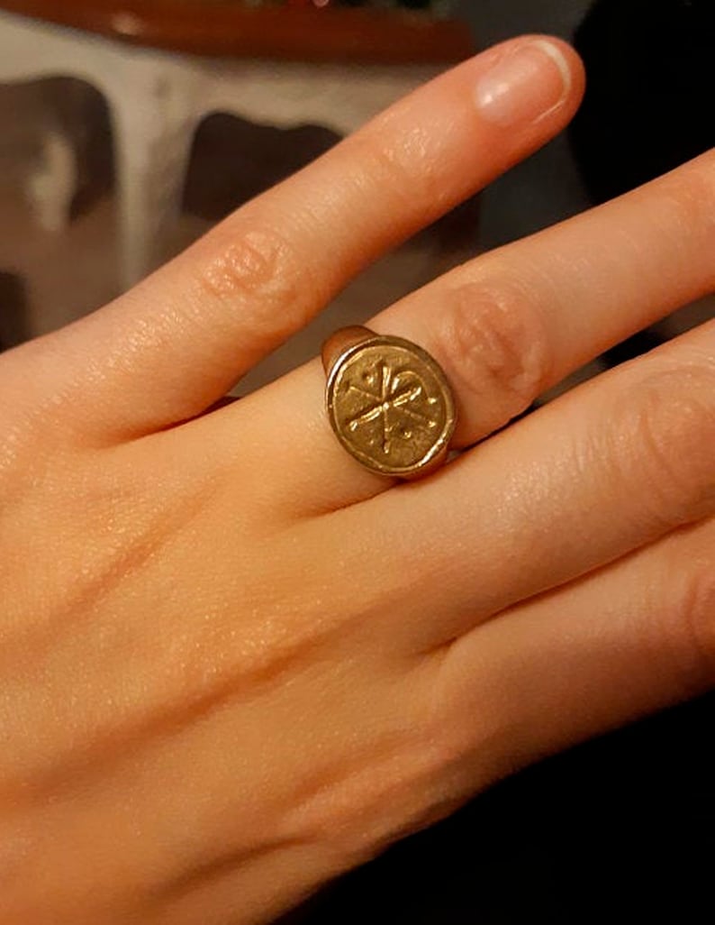 Puede incluir: Un anillo dorado con una cara circular que presenta una cruz y otros s&iacute;mbolos. El anillo se lleva en un dedo, con la mano en primer plano. El dise&ntilde;o del anillo es detallado y parece ser vintage o antiguo.