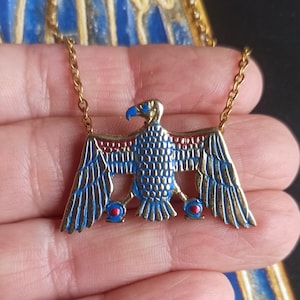 Könnte beinhalten: Eine goldene Kettenkette mit einem blauen und roten Emaille-Anhänger, der einen stilisierten ägyptischen Falken mit ausgebreiteten Flügeln darstellt.