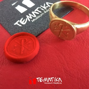 Puede incluir: Un anillo dorado con un emblema circular y un sello de cera rojo. El anillo y el sello est&aacute;n sobre una superficie roja, con una tarjeta negra con el texto "TEMATIKA Museum Replicas".