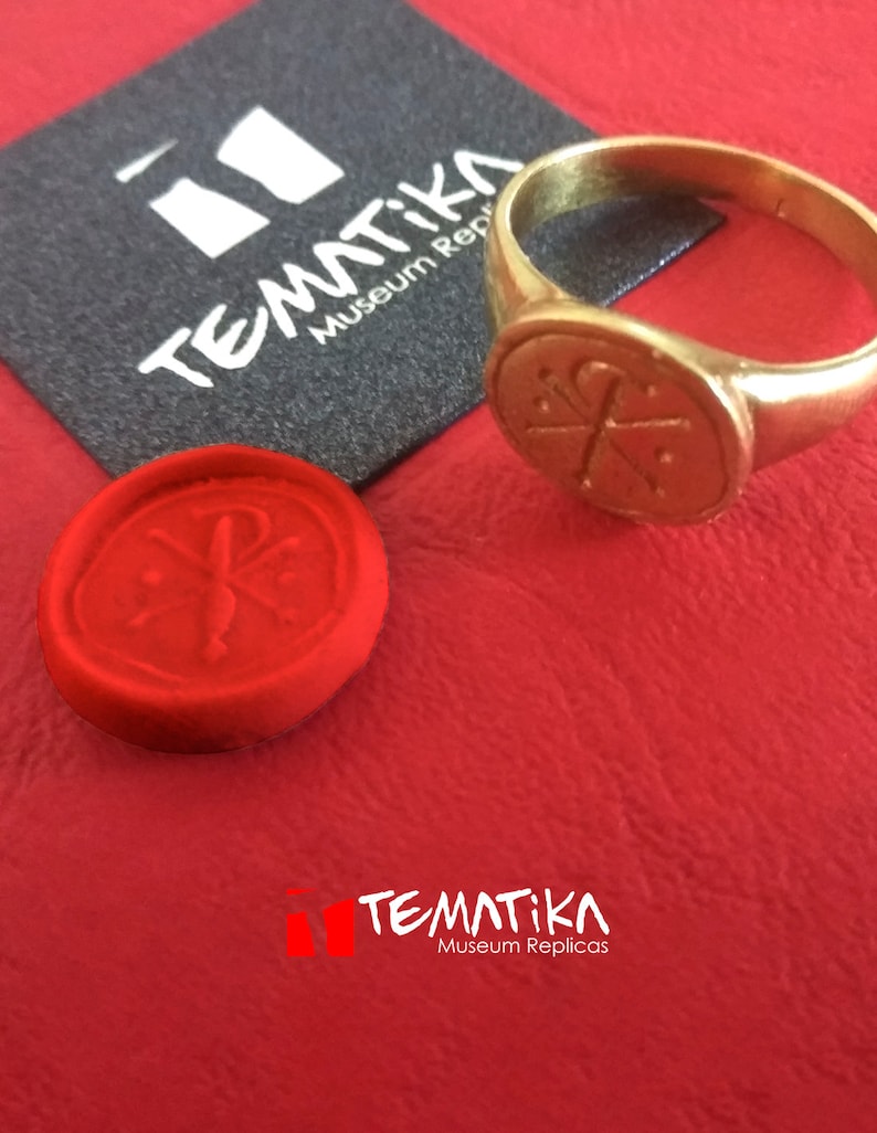 Puede incluir: Un anillo dorado con un emblema circular y un sello de cera rojo. El anillo y el sello est&aacute;n sobre una superficie roja, con una tarjeta negra con el texto "TEMATIKA Museum Replicas".