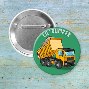 May include: A round button with a green background featuring a cartoon dump truck with the text "LIL' DUMPER" in white. The dump truck is yellow and black, with a bed full of brown material. The button is silver.