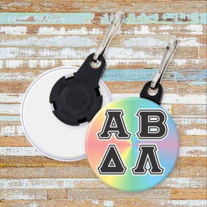 May include: Two round retractable badge reels. One is white with a black back, the other has a rainbow background with the letters AB and ΔΛ in black with a white outline. Both have a silver clip.
