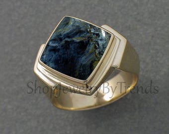 May include: A gold-toned ring with a square, dark blue gemstone. The gemstone has a marbled appearance with lighter blue and white streaks. The ring has a thick band and a modern design. The text "ShopJewelByTrends" is visible.