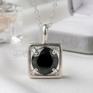 May include: A silver necklace featuring a square pendant frame that holds a round, faceted black gemstone in the center. The pendant is suspended from a delicate silver chain, showcasing a polished metallic finish and a secure prong setting.