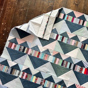 May include: A quilt made of denim and patterned fabric in shades of blue, pink, white, and green. The quilt is made up of triangles and is arranged in a chevron pattern. A paper pattern is visible on top of the quilt.