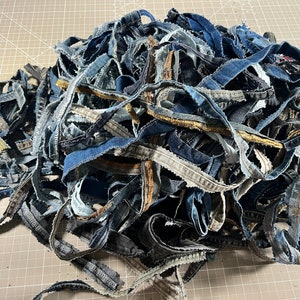 May include: A large pile of assorted denim fabric strips in various shades of blue, black, and grey. The strips feature visible seams, frayed edges, and contrast stitching, showcasing the raw texture of repurposed blue jean waistband material.