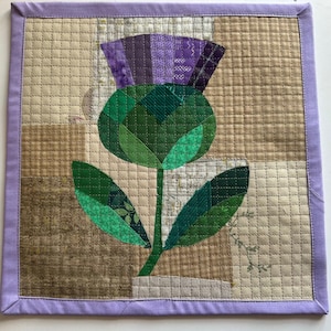 May include: A square quilted wall hanging featuring a stylized thistle. The thistle has a purple flower head and green leaves and stem, set against a patchwork background of beige and brown fabrics. The edges are finished with a lavender border.