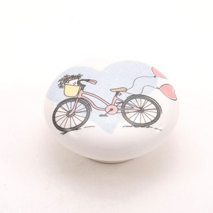 Könnte beinhalten: Ein weißer, herzförmiger Keramik-Knopf mit einem rosafarbenen Fahrrad mit einem Blumenkorb und zwei rosafarbenen Luftballons.