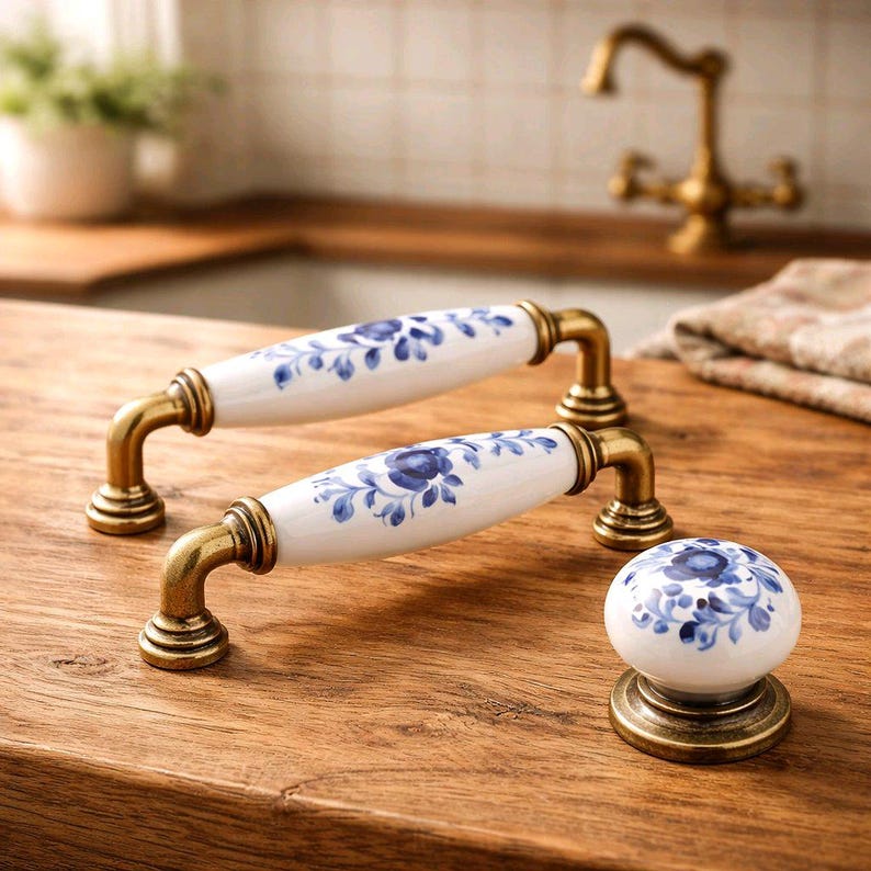 May include: Two elongated cabinet handles and one round cabinet knob, each featuring white ceramic centers with blue floral patterns and antique brass-toned metal bases, arranged on a wooden surface.