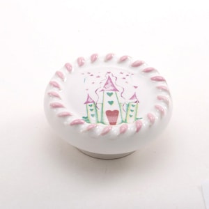 Op de afbeelding: Witte keramische knop met een roze en groen kasteelontwerp. Het kasteel heeft een roze dak en groene torens met roze harten. De knop is omgeven door een roze touwontwerp.