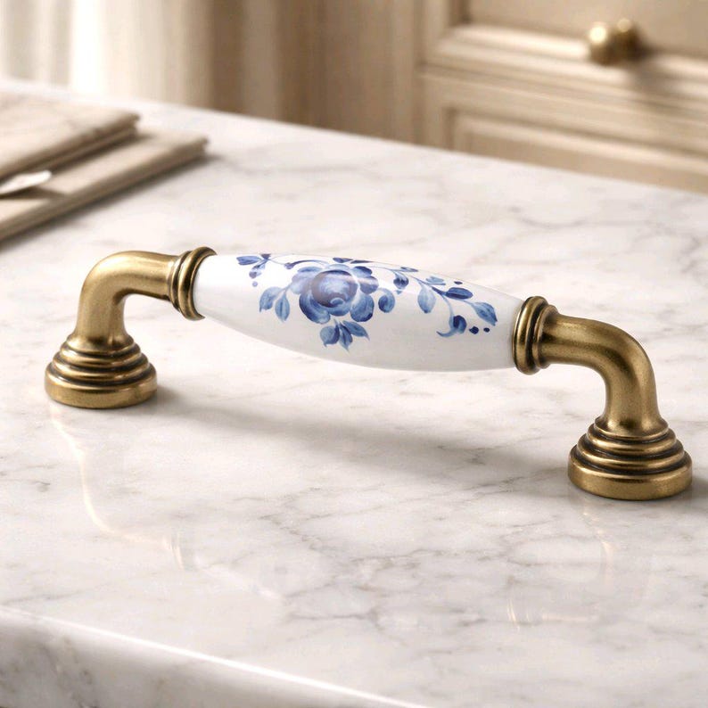 May include: A cabinet handle featuring a white ceramic center with a blue floral pattern and antique brass-toned metal ends. The handle has a curved, elongated shape with two circular mounting bases, showcasing a classic design and finish.