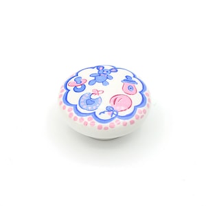 May include: White ceramic knob with a blue and pink baby-themed design. The design features a teddy bear, pacifiers, and a bottle.
