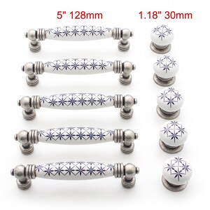 Könnte beinhalten: Fünf weiße Keramik-Schubladengriffe mit blauem Sternmuster und antikem silberfarbenem Metallbeschlag mit 128 mm sowie fünf passende runde Knöpfe mit 30 mm. Die Beschläge haben eine texturierte, verwitterte Metalloberfläche.