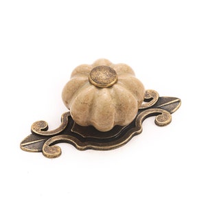 May include: A ceramic pumpkin-shaped knob with a gold-tone metal base and ornate scrollwork. The knob is beige with a marbled effect.