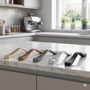 May include: Five curved cabinet pulls in various metallic finishes: gold, copper, gunmetal, silver, and black. The hardware is displayed on a marble countertop in a kitchen setting. These pulls are designed for cabinets and drawers.