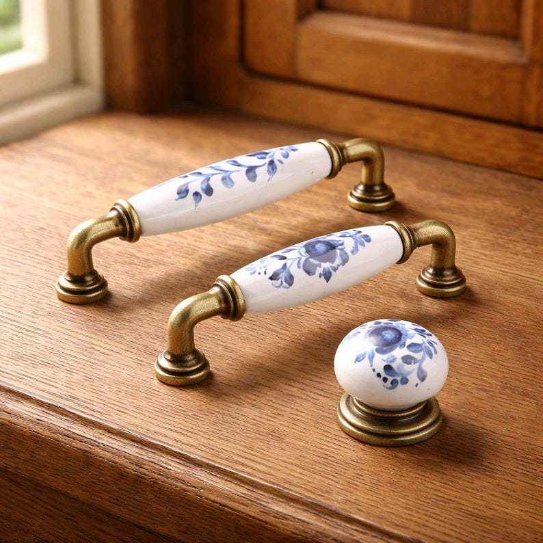 May include: Two elongated cabinet pulls and one round cabinet knob, each featuring white ceramic centers with blue floral and leaf patterns. The hardware components are connected by antique brass-toned metal bases and curved mounting brackets.