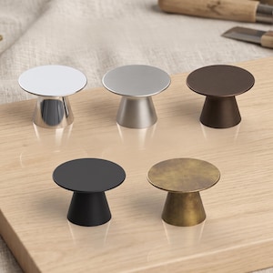 May include: Five circular cabinet knobs with a pedestal base, arranged in a group. The finishes include polished chrome, brushed nickel, dark bronze, matte black, and an aged brass texture, each featuring a flat top and a tapered conical stem.
