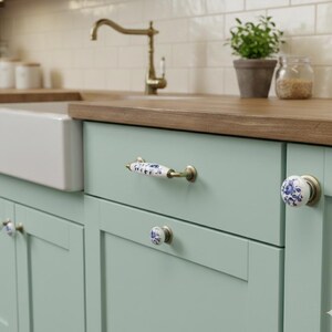 May include: A set of light green kitchen cabinets featuring white ceramic hardware with blue floral patterns. The hardware includes a horizontal drawer pull and two round knobs, all mounted with antique brass-toned metal fixtures.