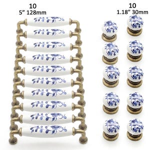 Op de afbeelding: Tien witte keramische ladehandgrepen met blauwe bloemmotieven en antiek messing beslag, 128 mm lang, samen met tien bijpassende ronde keramische knoppen met blauwe bloemmotieven, met een diameter van 30 mm.