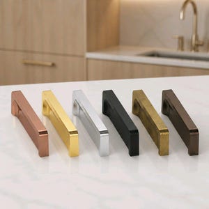 May include: Six cabinet pulls in various metallic finishes: copper, gold, silver, black, brass, and bronze. Each pull has a rectangular design with a curved handle, displayed on a white countertop. The background features a kitchen setting with wooden cabinets.