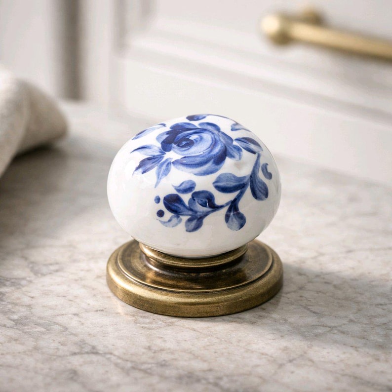 May include: A round ceramic cabinet knob featuring a white base with a hand-painted blue floral pattern. The knob is mounted on a circular, antique brass-toned metal base with a smooth, metallic finish and a slightly raised, stepped edge design.