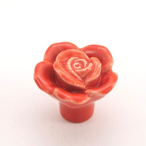 Op de afbeelding: Een keramische roosvormige kastknop in een fel oranje kleur. De roos heeft een gedetailleerd ontwerp met een centrale knop en bloemblaadjes.