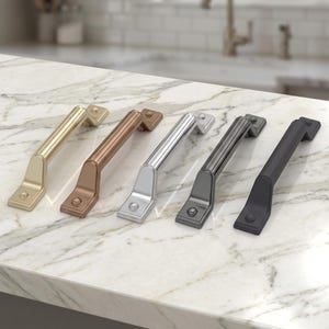 Può includere: Cinque maniglie per mobili in varie finiture metalliche: oro, rame, argento, canna di fucile e nero. Ogni maniglia ha una base rettangolare e un'impugnatura curva, che mostra una gamma di opzioni hardware per l'arredamento.