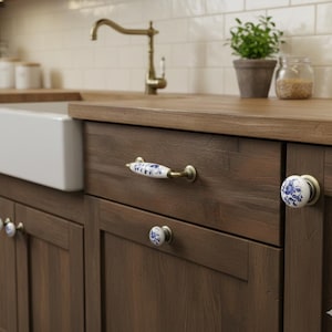 May include: A set of white ceramic cabinet knobs and a drawer pull featuring blue floral patterns and antique brass hardware. The hardware is mounted on dark brown wooden kitchen cabinets, including a long handle and several round knobs.