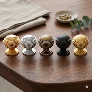 May include: Five decorative cabinet knobs in various finishes: gold, silver, bronze, black, and another gold. Each knob has a round, ribbed design. The knobs are arranged on a dark wooden surface, suggesting a home decor theme.