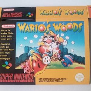 Puede incluir: Una caja de juego de Super Nintendo amarilla y azul con una imagen de Wario en la parte delantera. El juego se llama "Wario's Woods". La caja tiene texto en inglés y francés.