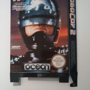 Op de afbeelding: Een vintage Nintendo Entertainment System game cartridge doos voor RoboCop 2. De doos is zwart met een rode rug en heeft een close-up afbeelding van RoboCop's helm en gezicht. De tekst "RoboCop 2" is in wit op de voorkant van de doos gedrukt. De doos bevat ook de logo's van Orion Pictures Corporation, Ocean Software en Nintendo.