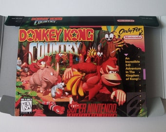 May include: A Donkey Kong Country Super Nintendo game box. The box art features Donkey Kong holding bananas, with other characters and the game title in a colorful design. The box is in a gray cardboard container.