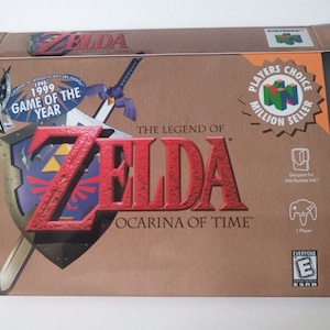 Peut inclure: Boîte de jeu The Legend of Zelda: Ocarina of Time pour Nintendo 64. La boîte est marron avec un logo Zelda rouge et or. La boîte a un autocollant Players Choice Million Seller et un autocollant du jeu de l'année 1999.