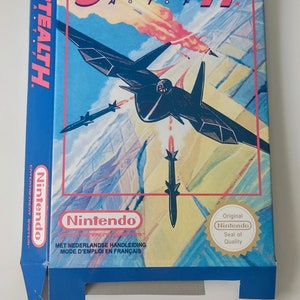 Op de afbeelding: Een blauwe en witte Nintendo-spelcartridgebox voor het spel "Stealth" met een zwarte straaljager die over een bergketen vliegt. De doos heeft een rood en wit "Nintendo"-logo en een "Keurmerk"-sticker.