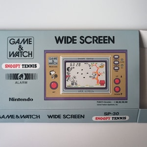 Puede incluir: Una consola de juegos portátil Game & Watch Wide Screen azul y blanca con un juego de Snoopy Tennis. La consola tiene un botón rojo y una pequeña pantalla con un juego en progreso. La caja está etiquetada como "SP-30" y "Snoopy Tennis".