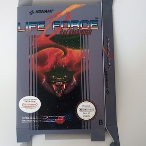 Pode incluir: Uma caixa de cartucho de jogo Nintendo cinza e preto para o jogo Life Force: Salamander, com uma criatura de dragão verde e vermelha sobre um fundo vermelho. A caixa tem o texto "KONAMI" e "LIFE FORCE" nela.