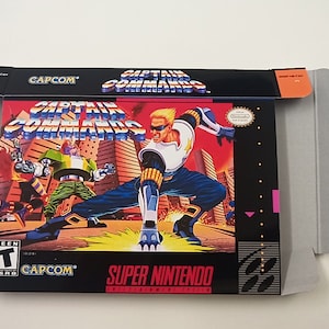 May include: A Super Nintendo game cartridge box for Captain Commando. The box is black with a red, yellow, and blue illustration of the game's characters. The text "CAPCOM" is printed in white on the box.