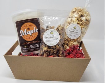 May include: A brown cardboard gift basket filled with red shredded paper. The basket contains a clear plastic container of white cotton candy with a brown label that says "Cotton Maple Candy", a clear plastic bag of maple candied nuts, and a clear plastic bag of maple popcorn. The labels on the bags of nuts and popcorn both say "Twin Hill Farm" and include the farm's address and phone number.