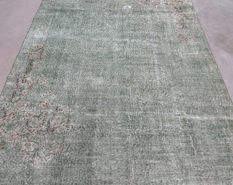 Puede incluir: Una alfombra rectangular en un tono verde salvia apagado, con una textura desgastada y envejecida. La alfombra muestra patrones florales intrincados y descoloridos en tonos marrones y crema suaves concentrados en las esquinas.