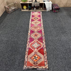 May include: A long, narrow runner rug featuring a vibrant magenta and red base with a series of repeating diamond-shaped medallions. The geometric patterns include intricate floral and star motifs in shades of orange, cream, and dark brown.