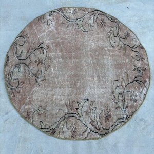 May include: A round rug featuring a faded dusty rose and beige color palette with a distressed texture. The design includes intricate black and dark brown floral and vine motifs arranged in a circular border pattern around the center.
