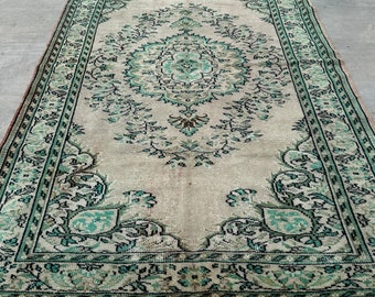Puede incluir: Una alfombra rectangular con un campo beige y un medallón central en tonos verde azulado, negro y marrón. La alfombra presenta un borde floral detallado y patrones de enredaderas intrincados, terminada con flecos blancos cortos.