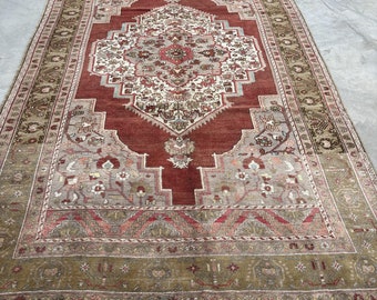 Puede incluir: Una alfombra rectangular que presenta un gran medallón central en forma de diamante en tonos crema y rojo. La alfombra muestra intrincados patrones florales y geométricos, con un borde ancho en verde oliva apagado y rojo.
