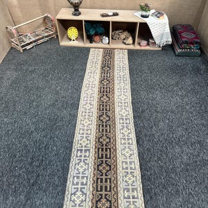 May include: A long, rectangular runner rug featuring a central dark brown panel with a repeating geometric pattern, flanked by two lighter beige side panels with intricate, symmetrical, multi-colored tribal-inspired motifs and borders.