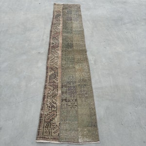 May include: A long, narrow rug with a geometric pattern in shades of brown, green, and beige. The rug has a fringe on both ends.