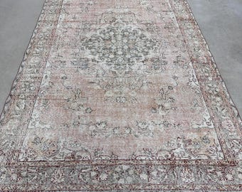 Puede incluir: Una alfombra rectangular con un fondo de color melocotón y rosa viejo descolorido, que presenta un medallón central y patrones florales intrincados en el borde en tonos gris carbón, crema y marrón apagado, con textura desgastada.