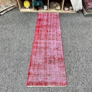 May include: A long, rectangular runner rug featuring a distressed, faded pattern in vibrant shades of red and magenta. The rug has a worn, vintage-inspired texture with darker, muted black and charcoal accents distributed across the surface.